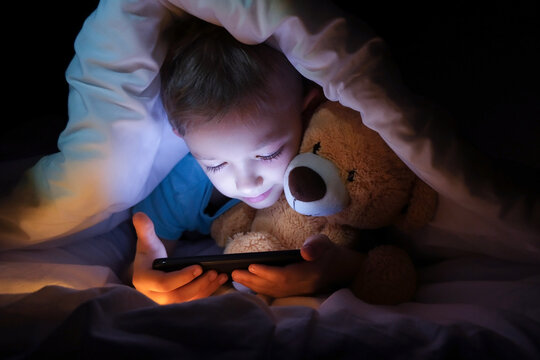 happy boy lies with toy bear in bed under a blanket and using a digital tablet smartphone device in the dark.