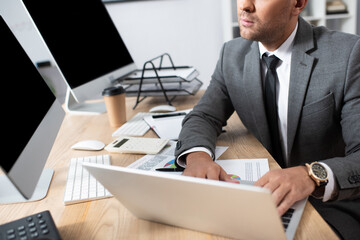 cropped view of trader typing on laptop near monitors with blank screen, blurred foreground