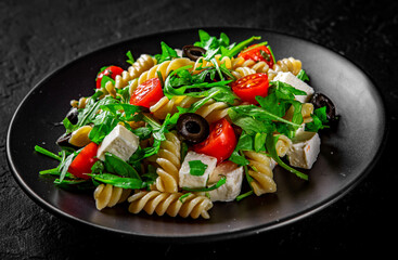 Salad with pasta, arugula, tomato, cheese and olives on black plate on Dark grey black slate background