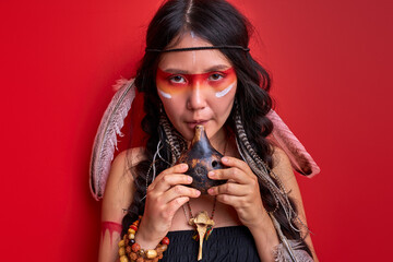 shaman woman using authentic indian whistle isolated on red background, female with feathers on head shamaning. portrait