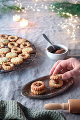 Christmas Linzer cookies filled on cooling rack, metal plate. Sandwich cookies filled with hazelnut spread, nougat cream. Hand holds cookie. Xmas garland on linen tablecloth. Natural light.