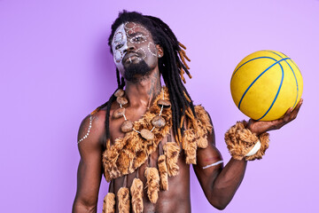 black pagan male with national make-up holding basketball ball in hands, wearing authentic traditional cape on naked body. purple background
