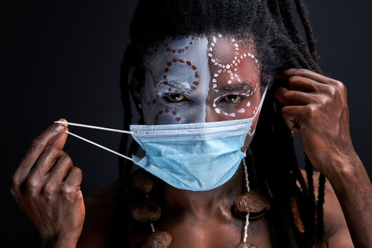 Aborigen Male Wearing Medical Mask On Face During Coronavirus, Covid-19 All Over The World. Man With Shamanic Paintings On Face Looks At Camera Isolated On Black Background