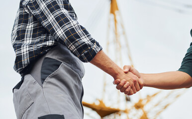 Making handshake. Two construction workers in uniform and safety equipment have job on building...