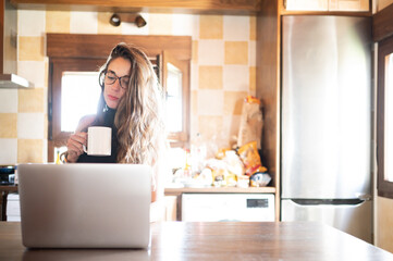 Beautiful and elegant businesswoman in front of her laptop while having a coffee in the kitchen of her house. New work reality