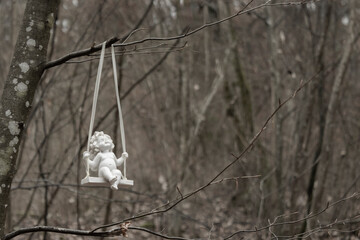black and white closeup of the figure of an angel sitting on a seesaw in the forest, with the cold bare trees in the background