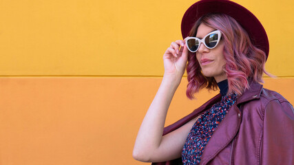 A lady in pink with sunglasses and a hat in front of a yellow wall. Negative space or copy space for text.