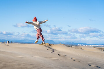 une enfant avec un bonnet rouge court les bras écartés en direction de la mer sur des dunes