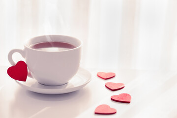 Fototapeta na wymiar Romantic tea party for lovers on Valentines day. Red heart shaped figurins with white cup of tea on light background. Good morning with hot tea concept. Space for text. tonned