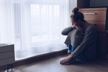 A devastated sad woman is sitting on the floor in her living room looking at the window. She is depressed.