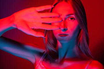 Unemotional young female covering eyes with hand in studio with red light