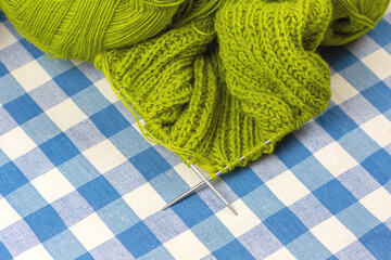 Knitting needles with green threads lie on a blue and white tablecloth.
