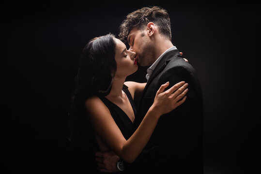 sexy woman with red lips kissing with man in suit isolated on black