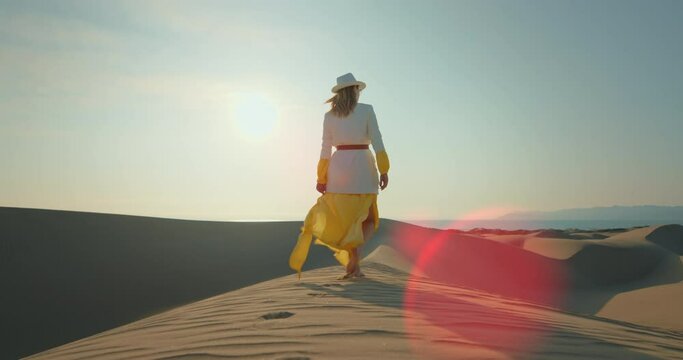 Young lady travelling in desert during safari trip. Back view of blonde woman in yellow dress walking barefoot on sand dune. Fashionable girl enjoying summer vacation in wild nature, 4k footage