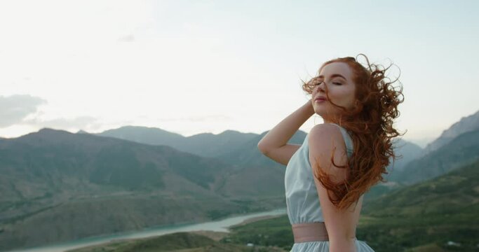 Gorgeous caucasian girl wearing a light dress is posing for a photo shoot on scenic mountain background while wind is blowing her red hair 4k footage