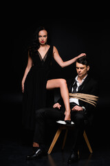 full length of dominant woman in dress pulling hair of tied submissive man sitting on chair on black