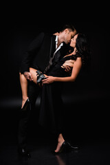full length of man lifting leg of dangerous and armed woman in dress and kissing her on black