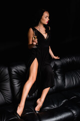 Fototapeta na wymiar brunette young woman in dress sitting on sofa and holding glass with champagne isolated on black