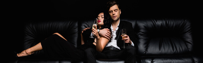 sexy couple holding glasses of champagne while sitting on sofa isolated on black, banner