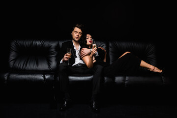 sexy couple holding glasses of champagne while sitting on sofa isolated on black