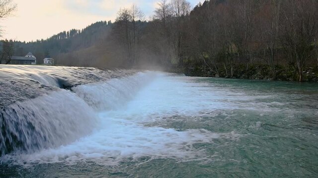 Pristine river flowing in amazing nature, Slovenia. Clean Sora river in Poljane valley. Cascading river with trees growing on river bank and houses in the distance. Static shot, real time