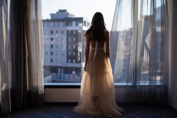 Portrait of gorgeous happy luxury stylish brunette bride in silk robe, standing at the window in skycraper. Beautiful details on wedding dress.  © Dragan