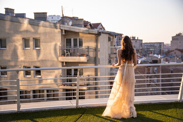 Portrait of gorgeous happy luxury stylish brunette bride in silk robe, standing on the top of skycraper, on the grass, watching the city at the sunset. © Dragan