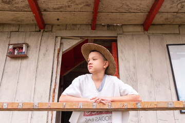 Niño campesino con sombrero parado en una casa de madera mirando