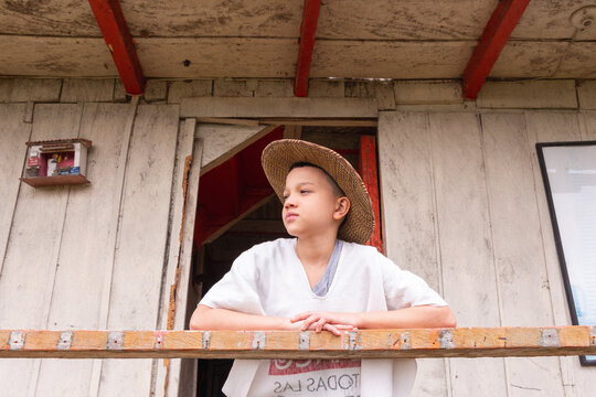 Ni&ntilde;o campesino con sombrero parado en una casa de madera mirando