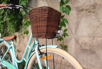 Vintage bicycle with wicker basket and headlamp stands near a concrete fence with green plants.