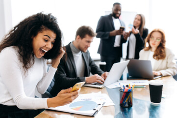 Joyful female employee sitting at the desk, looks at the phone and rejoices at luck. African american business woman reading good news, happy about successful project or startup, well done work