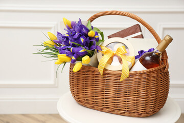 Wicker basket with gifts, bouquet and wine on white table indoors
