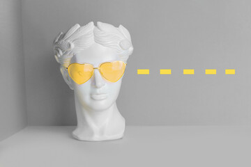 White sculpture of an antique head in yellow glasses with hearts. On a geometric background of two colors.