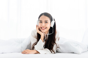 Beautiful young asian woman in headphones listening to music and lying down on bed at bedroom in morning time