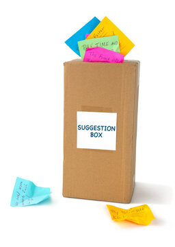 A tall cardboard SUGGESTIONS BOX with many colorful notes describing solutions to problems sticking out of the top of the drop slot and laying beside it.