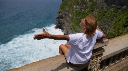 Middle aged woman sitting on a bench of a cliff spreads arms wide appreciating ocean view. Life after retirement