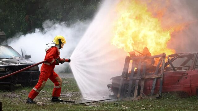 Brave firefighter in fire protection suit on safety rescue duty spray water from fire hose extinguishing crackle fire flames on explosion burning car. Fireman firefighting in danger training area