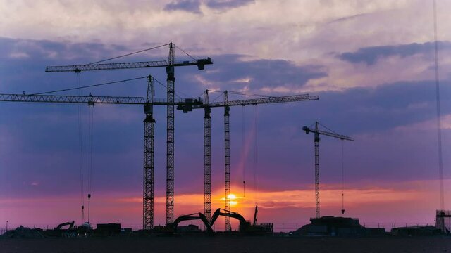 Silhouettes of high tower cranes operating with cargo near building at construction site under cloudy sky at sunset timelapse