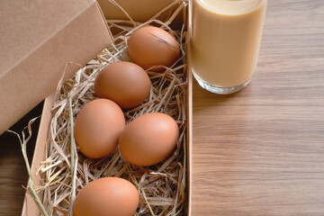 Eggs on straw in a brown box and a glass of milk placed on a wooden table.