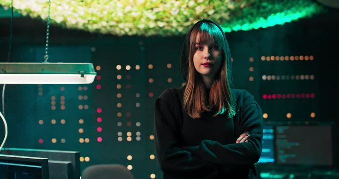 Portrait of a young female hacker looking at the camera in a secret hideout. In the background, monitors, cables and temporary servers.