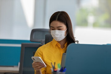 New normal of Asian woman in yellow shirt wearing surgical face mask using mobile phone at modern office or Co-working space