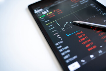 The pen points to the trader's selling price displayed as a graph falling on a tablet screen.