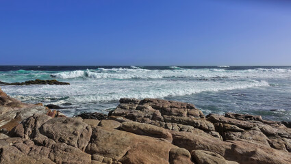 Fototapeta na wymiar The turquoise waves of the Atlantic Ocean leave a white foam on the coast. Ancient boulders in the foreground. Clear blue sky. Cape of Good Hope. South Africa