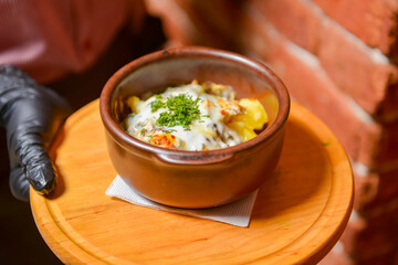Roasted vegetables and meat under melted cheese in a pot. Baked potatoes dish. Waiter serving dinner at restaurant.