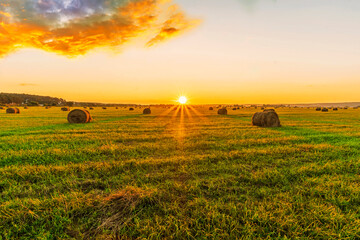 Scenic view at beautiful sunset in a green shiny field in willage farm with hay stacks, cloudy sky, golden sun rays, anazing summer valley evening landscape
