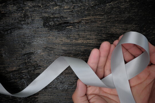 Top view of male hand holding gray ribbon on dark wood background with copy space. Brain cancer tumor awareness concept.