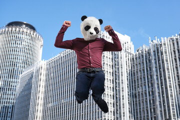 Young business man wearing a panda head mask, jumping in front of the corporate buildings. Bizarre businessman.