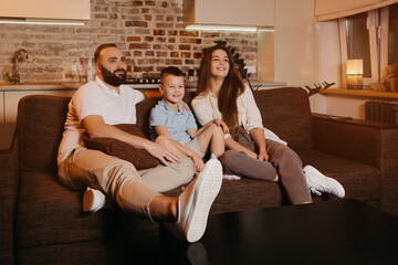 Dad with a beard, son, and young mom with long hair are watching with interest TV and smiling on the sofa in the apartment. The family is enjoying a happy evening at home.
