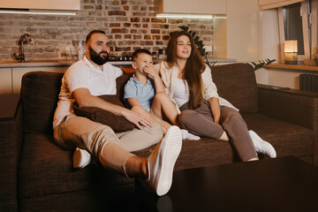 Dad with a beard, son, and young mom with long hair are watching TV and smiling on the sofa. The family is enjoying a happy evening at home. A child is laughing and covering his mouth with his hand.