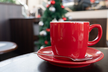 One lovely red coffee mug on the wooden table with the Christmas tree background. The aroma and tasty drink during the holiday afternoon at home. The caffeine beverage for breakfast.